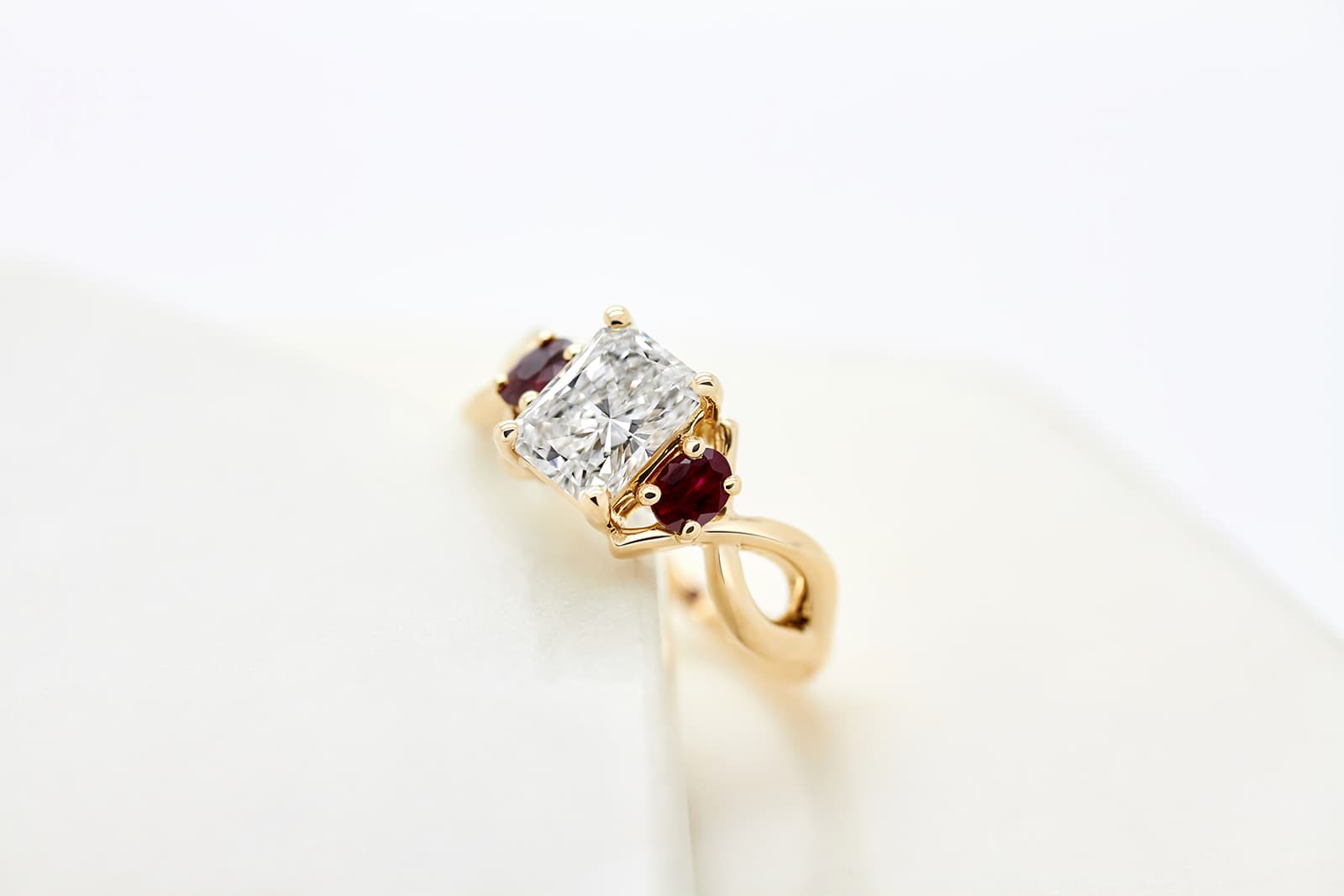  Taylor & Hart radiant-cut diamond engagement ring with oval-shaped ruby accent stones set in a twisted yellow gold band