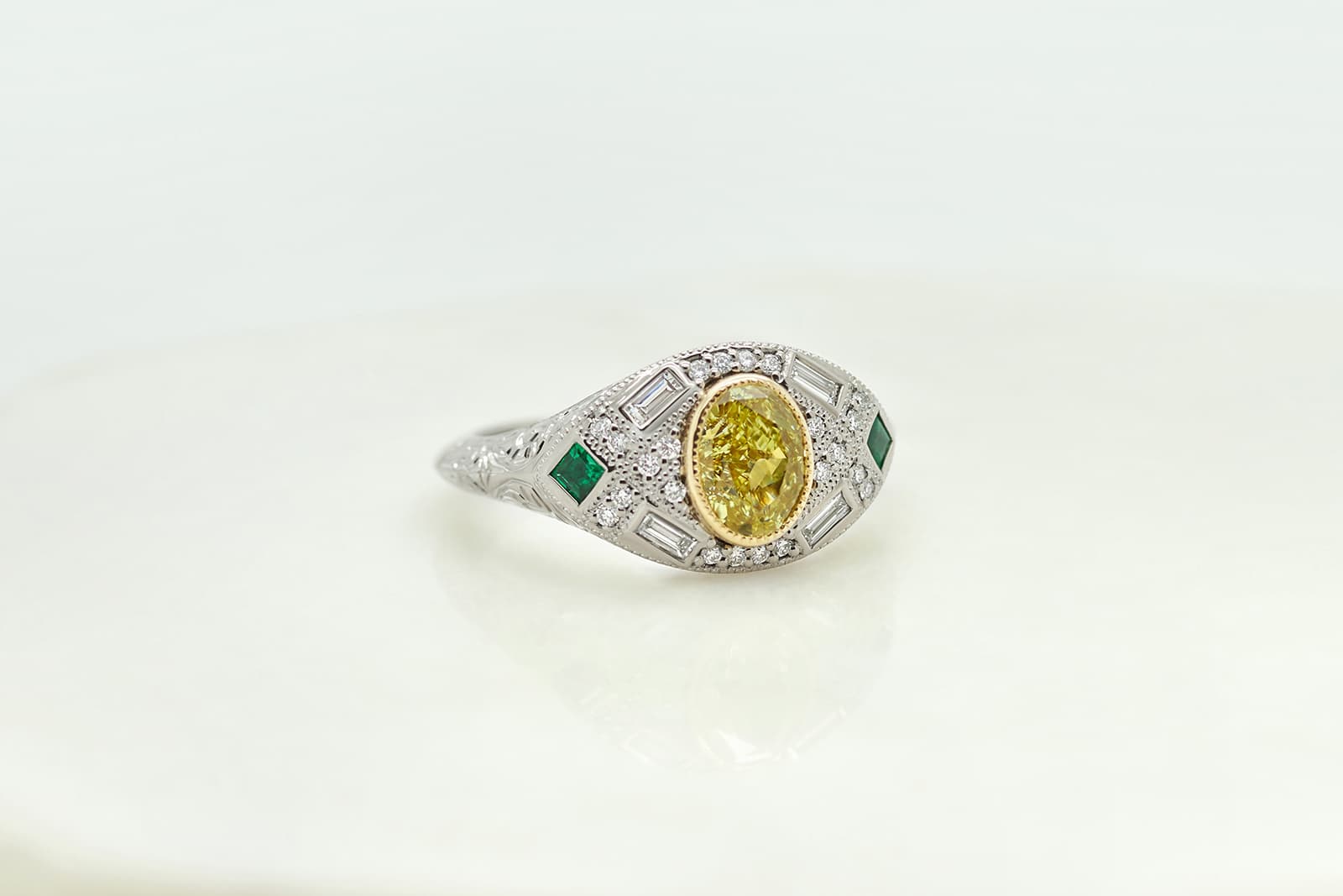  An Art Nouveau-inspired fancy intense yellow oval diamond engagement ring with round and baguette diamonds, princess cut emeralds, milgrain and engraving by Taylor & Hart