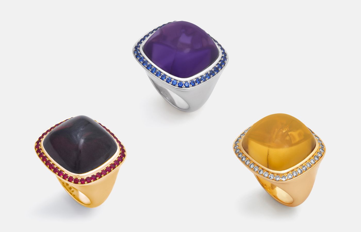 A trio of Magic Wish rings by Charlotte Reedtz Jewellery, including a garnet cabochon with ruby halo, an amethyst cabochon and blue sapphire halo, and a citrine cabochon and light brown diamond ring, all in 18k gold. 