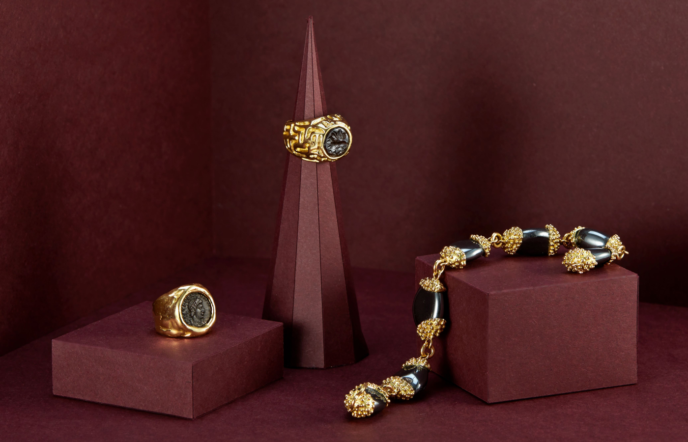  Elizabeth Gage Bronze Coin Molten Gold ring in gold and a bronze coin depicting Constans 337 - 350 AD, the Haematite Molten Gold ring in gold and a Sasanian haematite bead (ancient Persia) carved with an intaglio of a goat beneath a tree, 3rd - 6th Century AD, alongside the Hematite bracelet in gold and 7 hematite tumbled pebbles